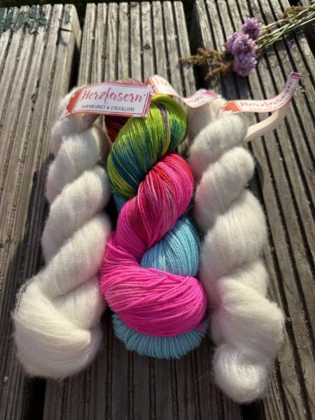 Handgefärbtes Garnset „Petit Provence“ von Herzfasern auf einer Holzbank im Sonnenlicht fotografiert. Das Set besteht aus einem bunten Strang Herzfasern No. 11 in der Farbe „Regenbogenkuss“ mit kräftigem Pink, Türkis, Grün und Violett sowie zwei fluffigen